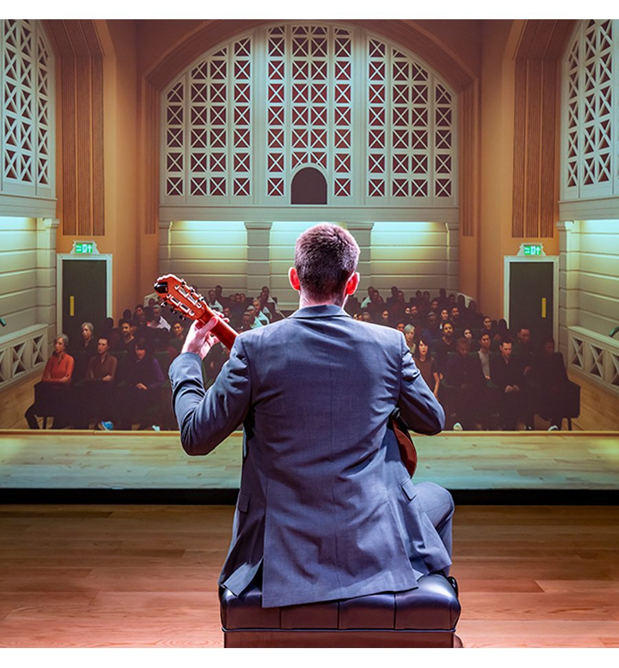 A male guitar student, wearing a smart suit, holding a guitar, performing to a virtual audience.